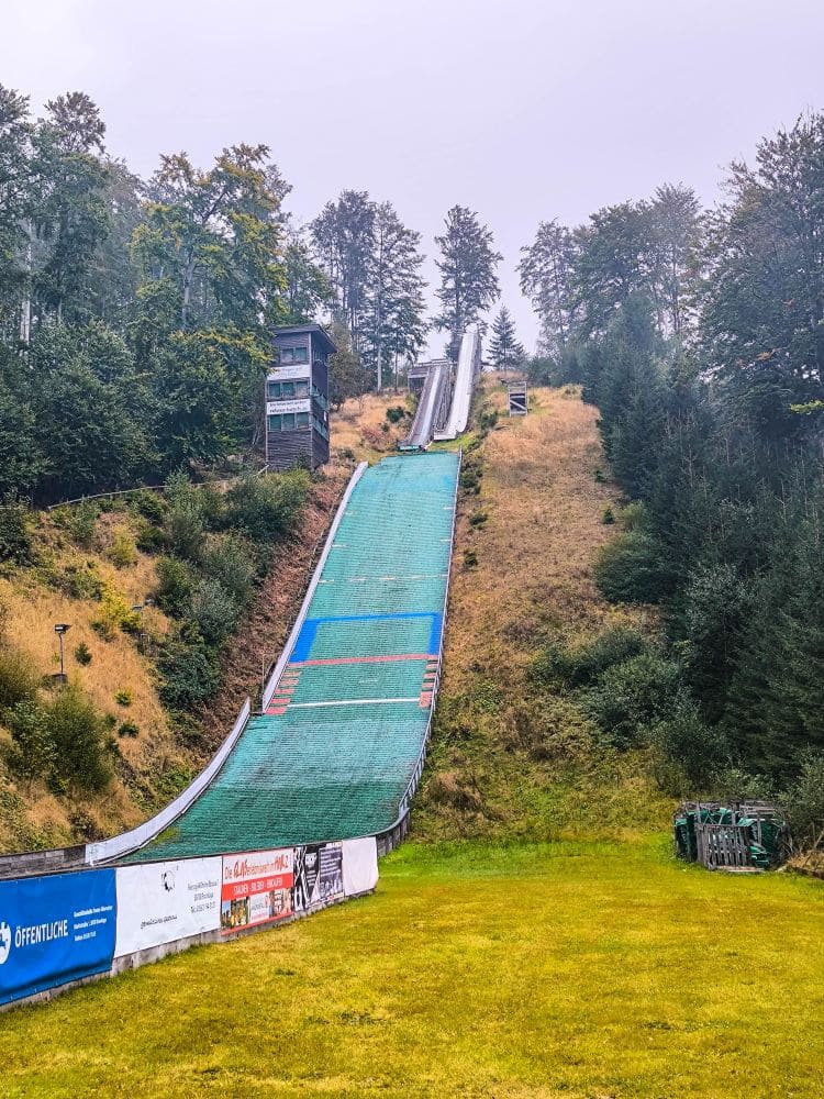 Eine große, mit Kunstrasen bedeckte Freiluft-Sprungschanze in Braunlage erhebt sich auf einem von Bäumen umgebenen Grashügel. Werbebanner säumen die Seiten, und links steht ein kleiner Aussichtsturm unter einem bedeckten Himmel.