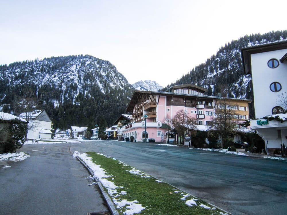 Ein ruhiges Bergdorf im Tannheimer Tal mit pastellfarbenen Gebäuden, einer breiten Straße und Schneeflecken. Im Hintergrund erheben sich schneebedeckte, mit Kiefern bewachsene Hügel und Berge unter einem blassen Himmel.