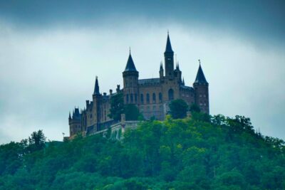 Eine große, mittelalterliche Burg mit mehreren spitzen Türmen, die an die Hohenzollern erinnert, thront auf einem grünen, bewaldeten Hügel unter einem wolkenverhangenen Himmel.