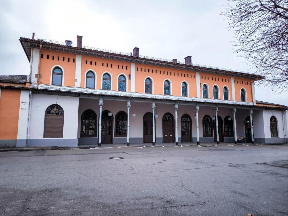 Ein zweistöckiges, orange-beiges Gebäude mit gewölbten Fenstern und Türen und einem überdachten, von Säulen getragenen Gang steht auf einer leeren, gepflasterten Fläche unter einem bewölkten Himmel in der Nähe des Starnberger Sees.