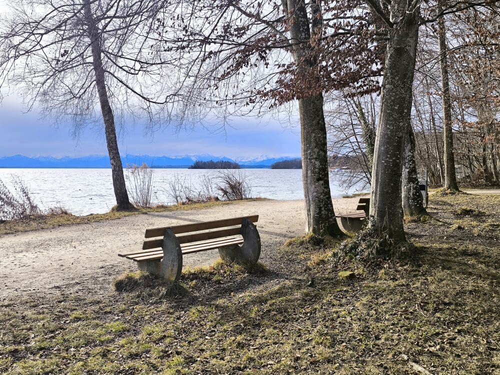 Zwei leere Holzbänke blicken auf das ruhige Wasser des Starnberger Sees, umgeben von blattlosen Bäumen. Der Himmel ist teilweise bewölkt, in der Ferne erheben sich die Berge, und die Szene strahlt eine ruhige, vorfrühlingshafte oder spätwinterliche Atmosphäre aus.