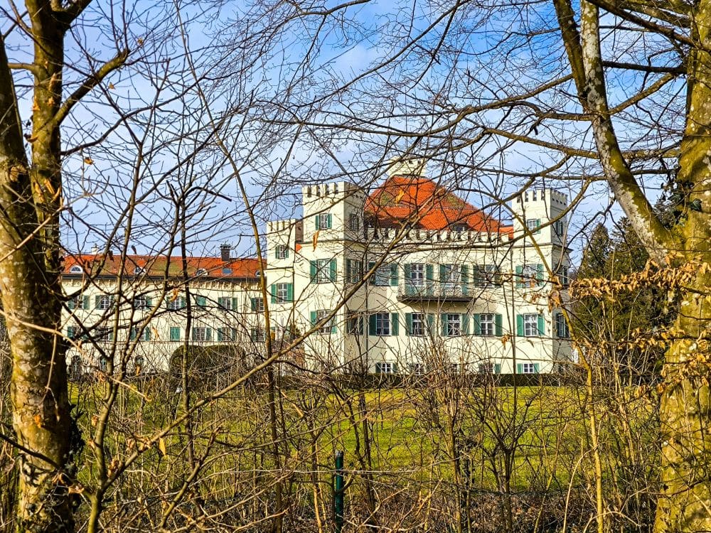 Ein großes weißes Gebäude mit rotem Ziegeldach und grünen Fensterläden steht in der Nähe des Starnberger Sees, gesehen hinter blattlosen Bäumen an einem sonnigen Tag, mit blauem Himmel und grünem Gras im Vordergrund.