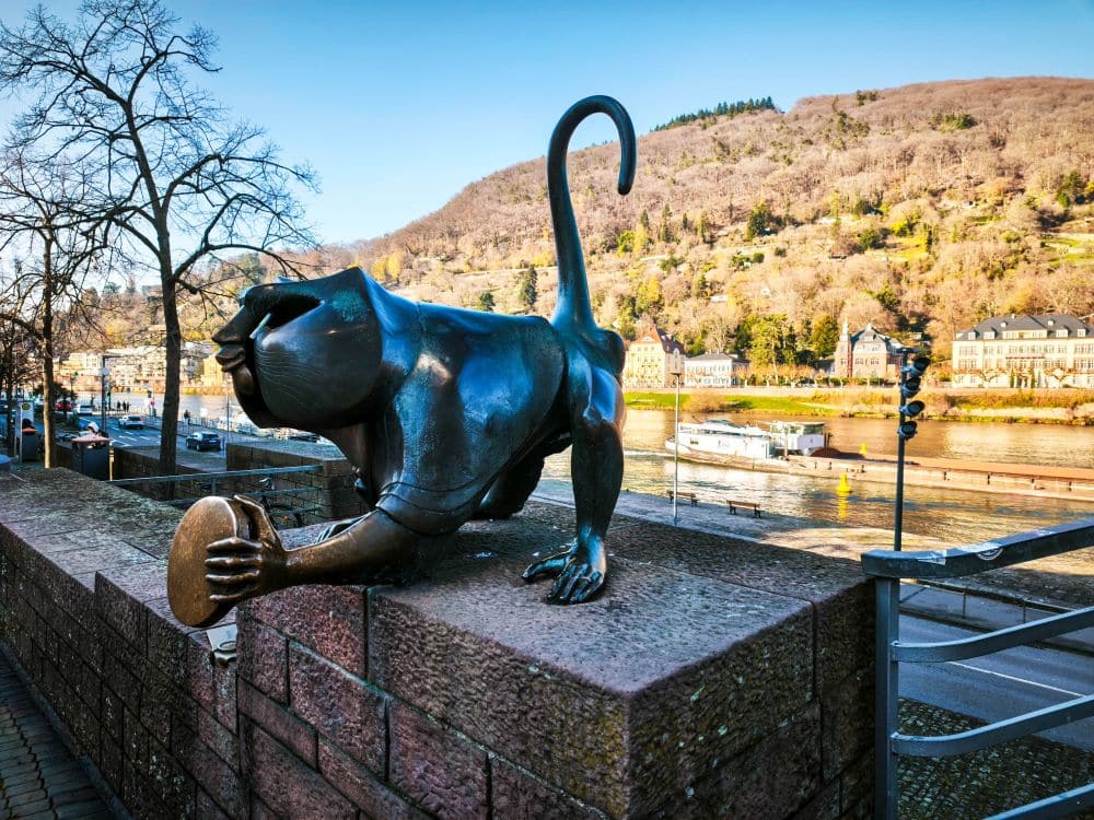 Eine bronzene Affenstatue mit hohlem Kopf und erhobenem Schwanz steht auf einer Steinmauer in Heidelberg und blickt auf den Fluss. Vor dem Hintergrund von Hügeln und historischen Gebäuden hält die Statue einen runden Gegenstand in einer Hand.