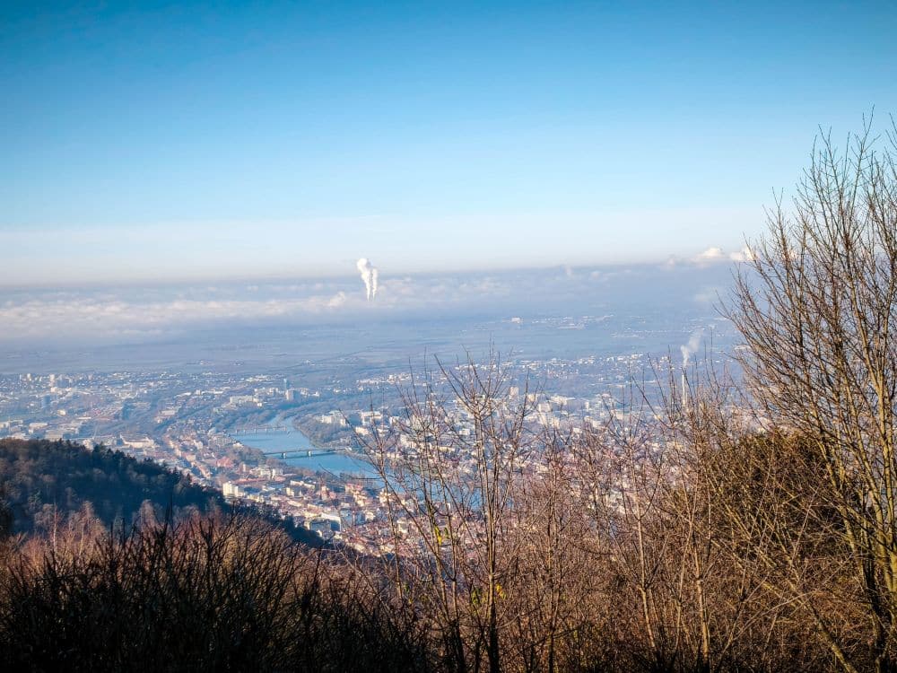 Blick von einer Anhöhe über Heidelberg mit verstreuten Gebäuden, Bäumen im Vordergrund, blauem Himmel und einer Rauch- oder Dampfwolke, die in der Ferne hinter einer Wolkenschicht aufsteigt.