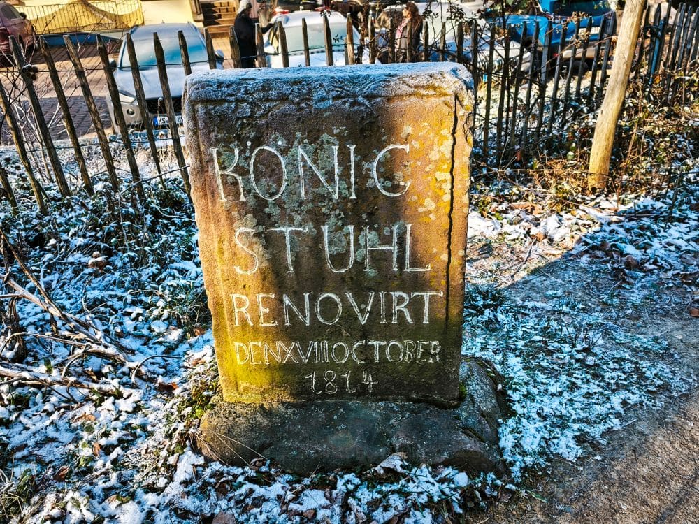 Ein verwitterter Gedenkstein mit weißer Schrift steht neben einem schneebedeckten Weg und einem Metallzaun in der Nähe von Heidelberg. Die Inschrift lautet "KÖNIG STUHL RENOVIRT DEN VII. OKTOBER 1814". Gefallene Blätter und leichter Schnee umgeben den Stein.