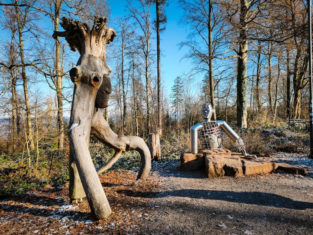 Eine hölzerne Skulptur, die einem Baum mit Beinen ähnelt, steht neben einer humanoiden Figur aus Metall, die auf einem Stein sitzt. Sie steht in einem Waldgebiet in der Nähe von Heidelberg, umgeben von hohen Bäumen und blauem Himmel.