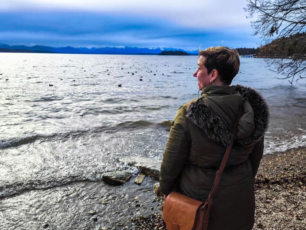 Eine Person in einem grünen Mantel mit einer braunen Umhängetasche steht am felsigen Ufer des Starnberger Sees, blickt über das Wasser auf die fernen Berge und beobachtet Vögel, die unter einem wolkenverhangenen Himmel über den See gleiten.