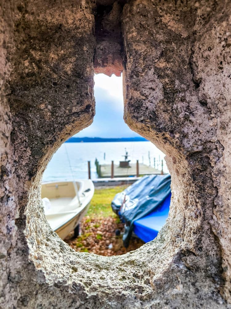 Blick auf ein kleines Boot und ruhiges Wasser durch eine runde, verwitterte Steinöffnung am Starnberger See, wobei sich das Sonnenlicht auf dem See spiegelt und eine blaue Plane ein Objekt in der Nähe abdeckt.
