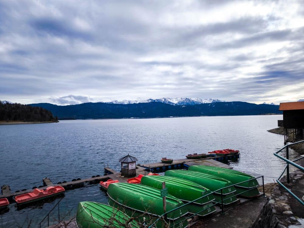Eine ruhige Szene am Walchensee: Eine Anlegestelle mit grünen Paddelbooten und roten Tretbooten ruht vor schneebedeckten Bergen und einem bewölkten Himmel. Sträucher säumen den Vordergrund und unterstreichen die friedliche Atmosphäre am See.
