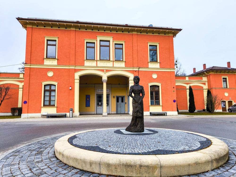 Eine Frauenstatue steht auf einem runden Steinpodest in der Nähe des Starnberger Sees, vor einem orangefarbenen Gebäude mit gewölbten Fenstern und Türen, umgeben von einer gepflasterten Fläche und einigen Bäumen.