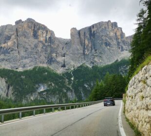 Ein Auto fährt eine kurvenreiche Bergstraße entlang, die von einer Steinmauer und dichten Bäumen gesäumt ist. Hoch aufragende Felsen und grüne, bewaldete Hänge ragen dramatisch in die Höhe - die atemberaubende Schönheit der alpinen Pässe.