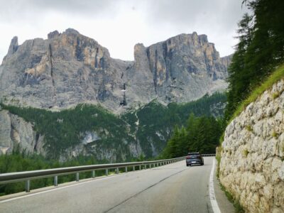 Ein Auto fährt eine kurvenreiche Bergstraße entlang, die von einer Steinmauer und dichten Bäumen gesäumt ist. Hoch aufragende Felsen und grüne, bewaldete Hänge ragen dramatisch in die Höhe - die atemberaubende Schönheit der alpinen Pässe.
