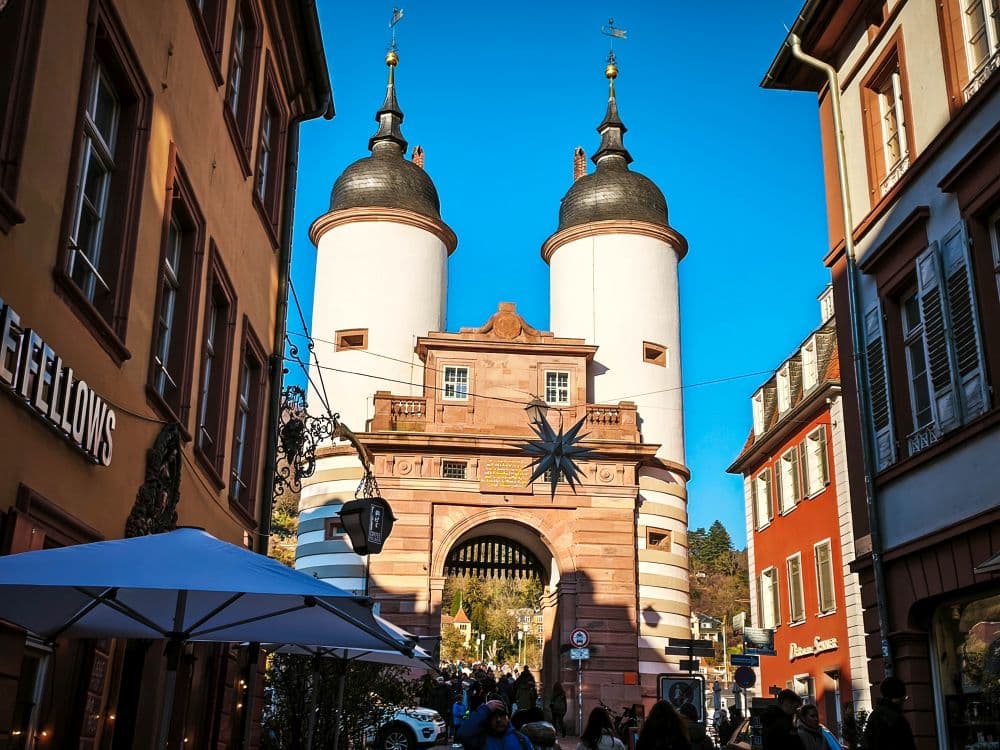An einem sonnigen Tag spazieren die Menschen in Heidelberg auf das historische Alte Brückentor mit seinen weißen Zwillingstürmen und schwarzen Kuppeln zu, umgeben von farbenfrohen Gebäuden und lebhaften Geschäften.