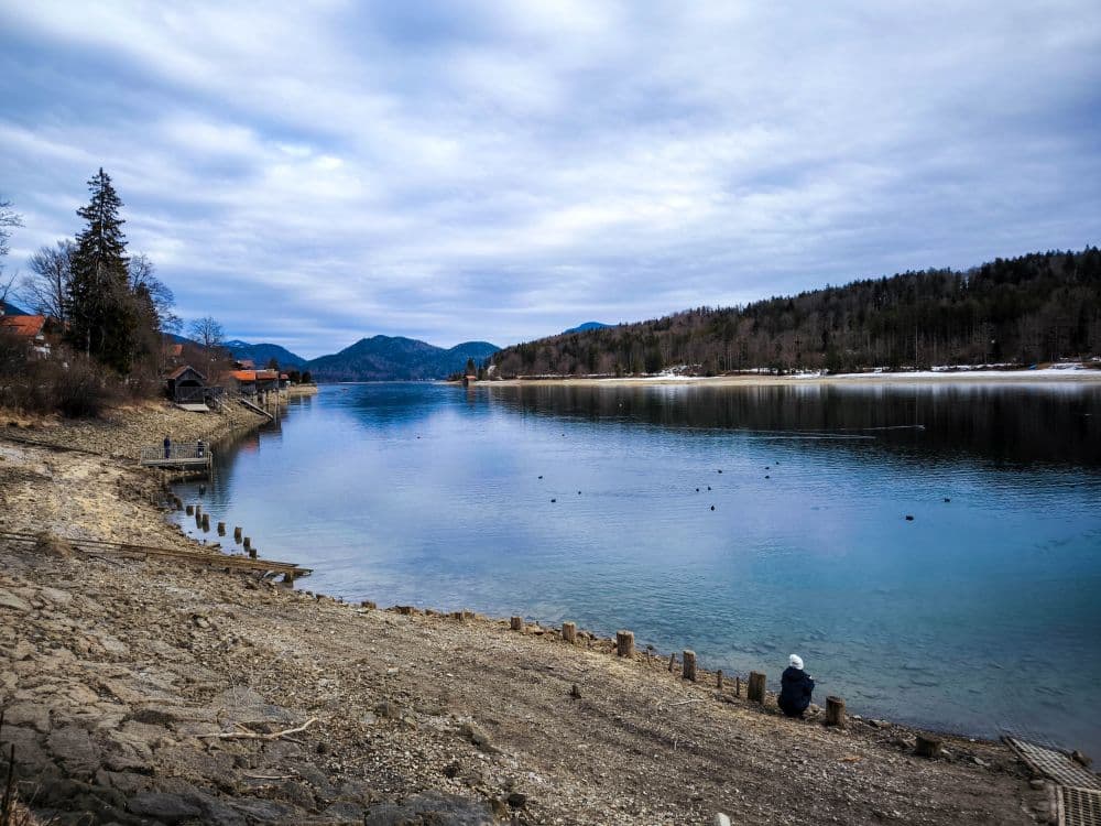 Ein ruhiger See, der Walchensee, gesäumt von einem felsigen Ufer und bewaldeten Hügeln unter einem bewölkten Himmel. Eine Person sitzt in der Nähe des Ufers, während eine andere auf einem kleinen Steg steht. In der Ferne sind Berge und verstreute Häuser zu sehen.