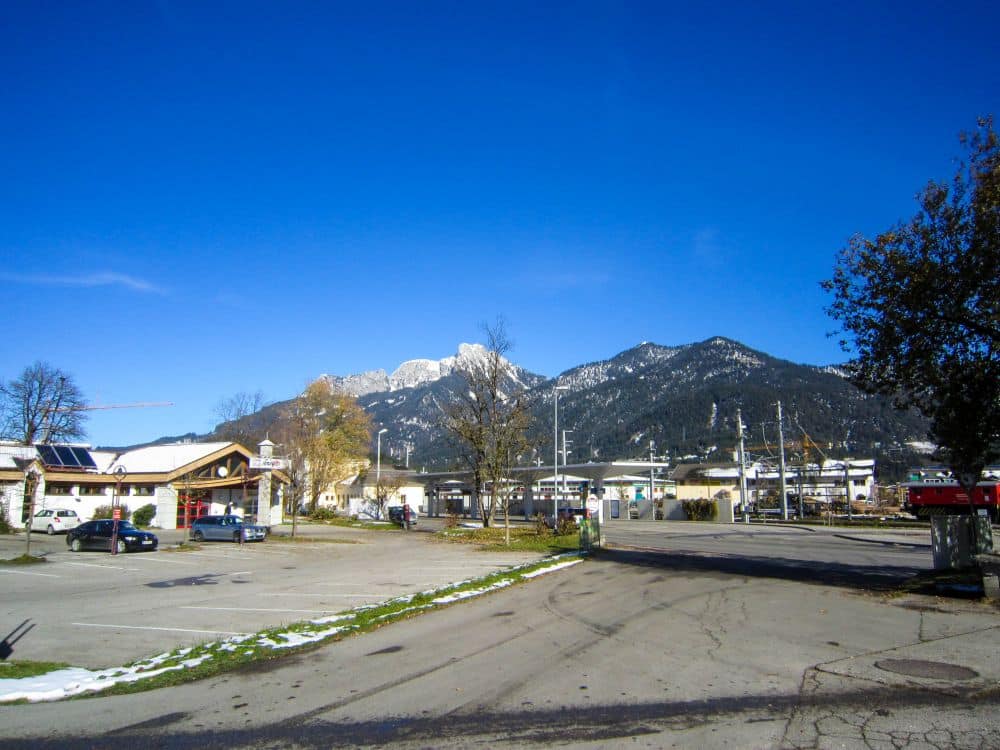 Ein größtenteils leerer Parkplatz mit ein paar geparkten Autos, kleinen Gebäuden und blattlosen Bäumen vor der atemberaubenden Kulisse der schneebedeckten Berge und des klaren blauen Himmels im Tannheimer Tal.