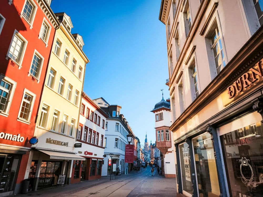 Ein heller, sonniger Tag in einer Heidelberger Straße, die von bunten Gebäuden, Geschäften und Cafés gesäumt ist. Die Straße ist ruhig, keine Menschen sind zu sehen, und der Himmel ist klar und blau.