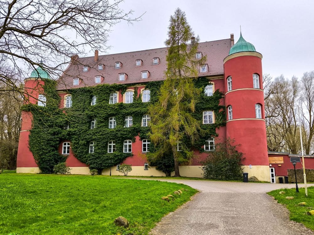 Ein großes rotes Gebäude auf Rügen mit grünem Wein, der einen Teil der Fassade bedeckt, runden Türmen an jeder Ecke und einem steilen Ziegeldach, inmitten einer Grünfläche mit Bäumen und einem Kiesweg, der zum Eingang führt.