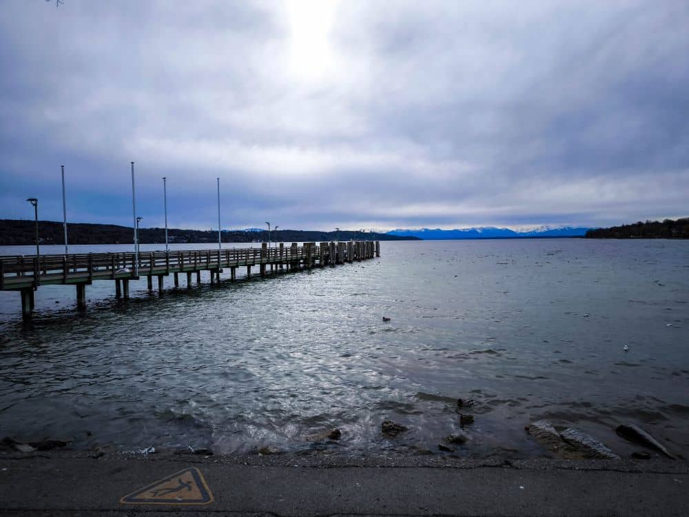 Ein langer Holzsteg ragt in das ruhige Wasser des Starnberger Sees unter einem wolkenverhangenen Himmel, mit schneebedeckten Bergen in der Ferne. Am felsigen Ufer steht ein Warnschild in der Nähe der Wasserkante.