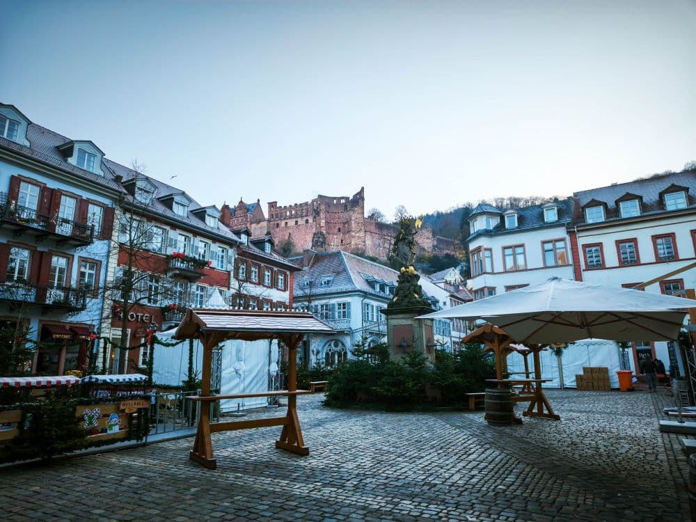 Ein kopfsteingepflasterter Platz in Heidelberg mit leeren Marktständen und einer Statue ist von historischen Gebäuden umgeben; ein großes Schloss thront auf einem Hügel im Hintergrund unter einem klaren Himmel.