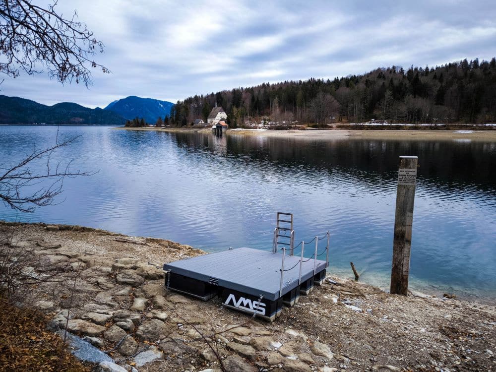 Ein kleiner Steg mit Geländer liegt am felsigen Ufer des Walchensees. Im ruhigen Wasser spiegeln sich ein bewölkter Himmel und bewaldete Hügel, während auf der anderen Seite des Sees Gebäude und Bäume zu sehen sind.