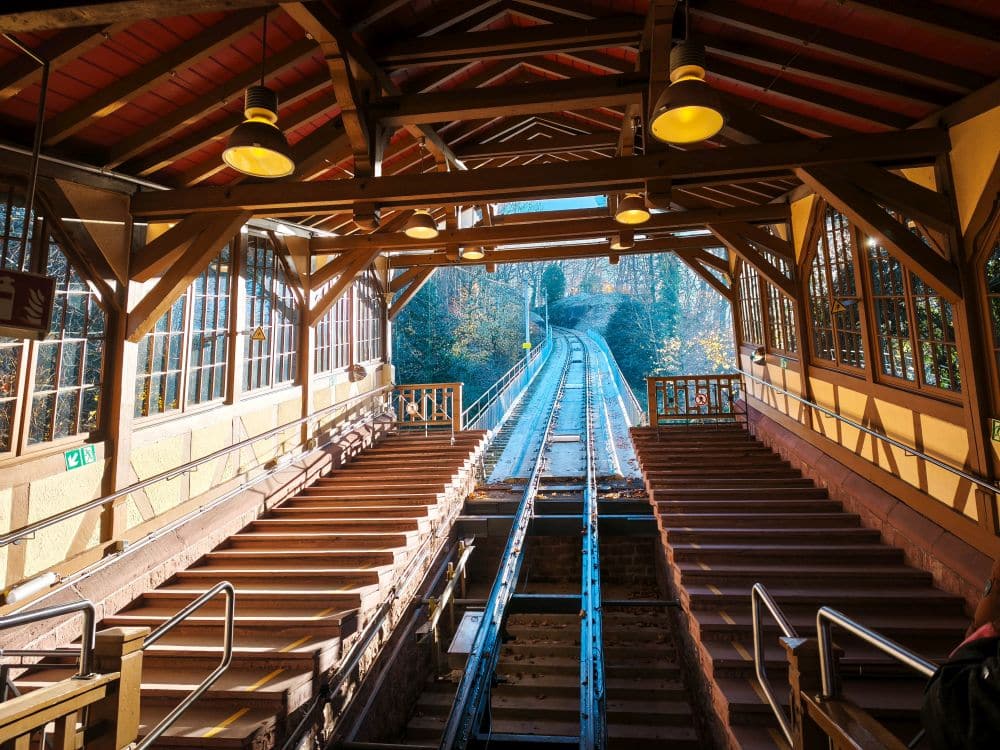 Eine überdachte Standseilbahn in Heidelberg mit zwei Holztreppen auf beiden Seiten der Gleise, die ins Sonnenlicht führen und einen Blick auf die Gleise bieten, die einen mit Herbstbäumen gesäumten Hügel hinaufführen.