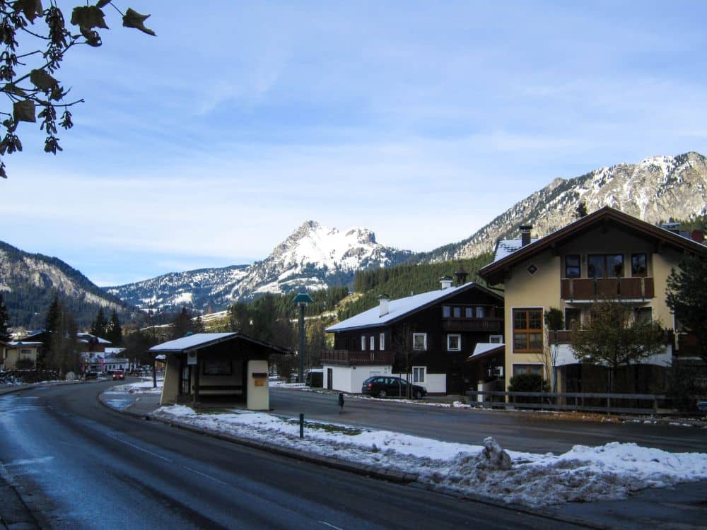 Ein ruhiges Bergdorf im Tannheimer Tal mit schneebedeckten Gehwegen, Holzhäusern und einer Bushaltestelle vor der Kulisse hoher, schneebedeckter Berge unter einem teilweise bewölkten Himmel.