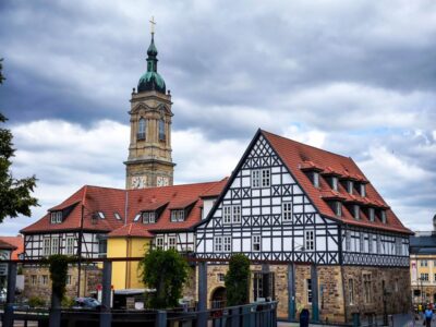 Ein historisches Fachwerkhaus mit steilem rotem Dach steht vor dem hohen Kirchturm von Eisenach mit Uhr und grüner Kuppel unter einem bewölkten Himmel. Die Szene zeigt den klassischen europäischen Baustil.