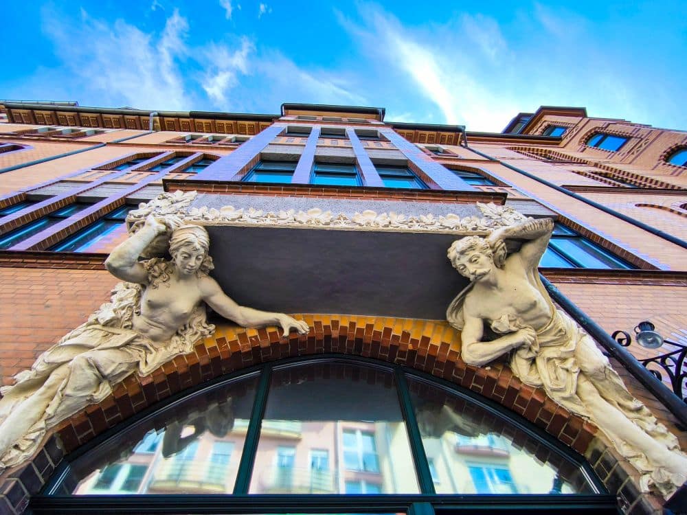 Tiefblick auf eine verzierte Eisenacher Gebäudefassade mit zwei großen Figuren, die ein dekoratives Gesims über einem Rundbogenfenster tragen, vor einem strahlend blauen Himmel mit Wolkenfetzen.