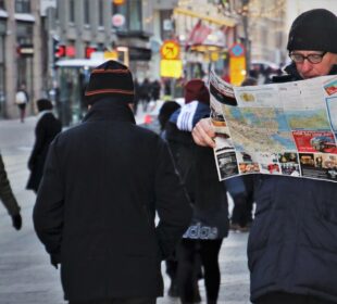 Ein Mann mit dunklem Mantel und Hut liest auf einer belebten Straße in Helsinki im Winter einen Stadtplan, während die Menschen gebündelt vorbeigehen. Im Hintergrund leuchten Gebäude und Lichter, perfekt für diejenigen, die die Stadt zu Fuß erkunden wollen.