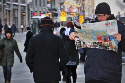Ein Mann mit dunklem Mantel und Hut liest auf einer belebten Straße in Helsinki im Winter einen Stadtplan, während die Menschen gebündelt vorbeigehen. Im Hintergrund leuchten Gebäude und Lichter, perfekt für diejenigen, die die Stadt zu Fuß erkunden wollen.