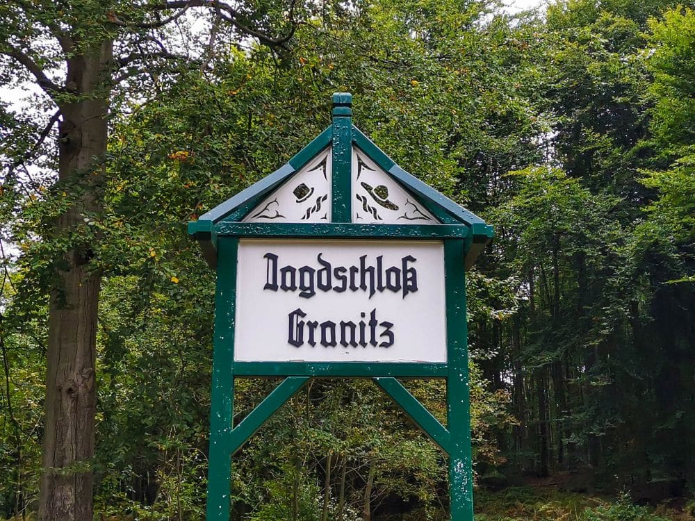 Zwischen hohen, dichten Bäumen in einem Waldgebiet auf Rügen steht ein hölzernes Schild mit grünem Rahmen, auf dem in schwarzen Buchstaben Jagdschloss Granitz steht.