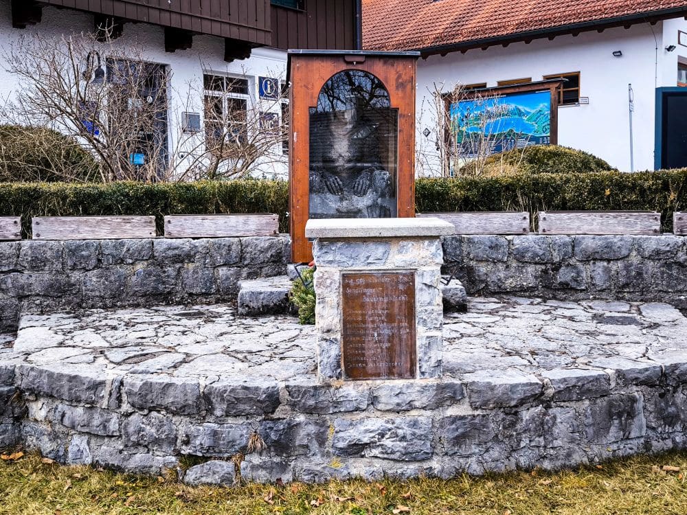 In der Nähe des Walchensees, vor weißen Gebäuden mit braunen Dächern, steht auf einer kreisförmigen gepflasterten Fläche, umgeben von niedrigen Steinmauern und Sträuchern, ein kleines Steindenkmal mit Gedenktafeln.