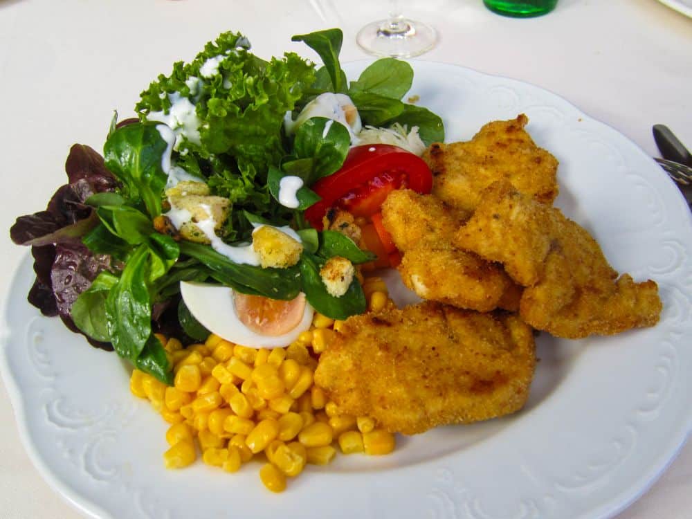 Ein weißer Teller mit panierten Hähnchenstreifen, grünem Blattsalat mit Dressing, Tomatenscheiben, einem halben gekochten Ei, Croutons und Mais erinnert an die frischen Aromen, die man nach einer Wanderung im malerischen Tannheimer Tal genießen würde.