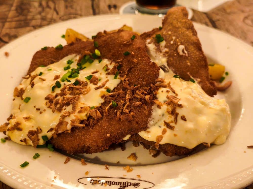 Ein Teller mit panierten und gebratenen Schnitzeln nach Heidelberger Art, belegt mit Sahnesoße, Schnittlauch und knusprigen Röstzwiebeln, serviert mit gerösteten Kartoffelstückchen auf einem weißen Teller.