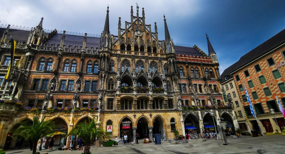 Ein prächtiges Gebäude im gotischen Stil mit verzierten Türmen und Bögen dominiert einen öffentlichen Platz in München, auf dem Menschen spazieren gehen und Palmen zu sehen sind. Der dramatische, teilweise bewölkte Himmel unterstreicht die Szene - perfekt, um sie mit Ihrer München City Card zu erkunden.