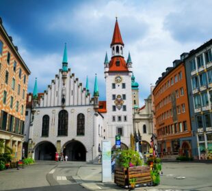 Ein lebendiger Stadtplatz in München, Deutschland, mit dem Alten Rathaus mit seinem hohen Uhrenturm, den bunten Türmen und den umliegenden historischen Gebäuden unter einem teilweise bewölkten Himmel.