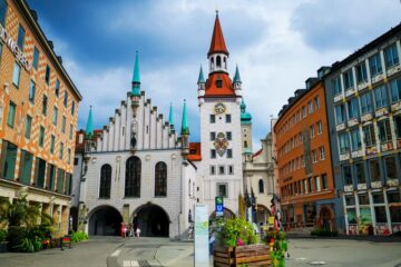 Ein lebendiger Stadtplatz in München, Deutschland, mit dem Alten Rathaus mit seinem hohen Uhrenturm, den bunten Türmen und den umliegenden historischen Gebäuden unter einem teilweise bewölkten Himmel.