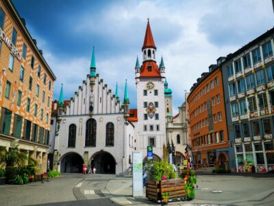 Ein lebendiger Stadtplatz in München, Deutschland, mit dem Alten Rathaus mit seinem hohen Uhrenturm, den bunten Türmen und den umliegenden historischen Gebäuden unter einem teilweise bewölkten Himmel.