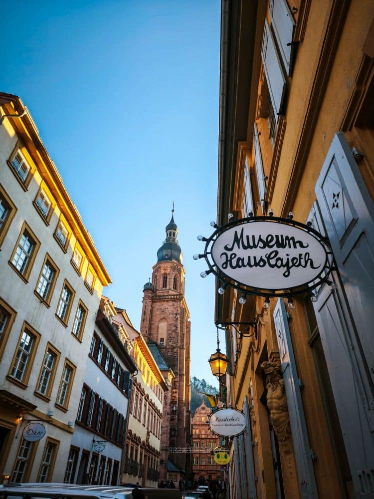 Eine schmale Straße in Heidelberg schlängelt sich an gelben und weißen Häusern vorbei. Im Hintergrund erhebt sich ein Schild für das Museum Haus Cajeth und ein hoher Kirchturm unter einem klaren blauen Himmel.