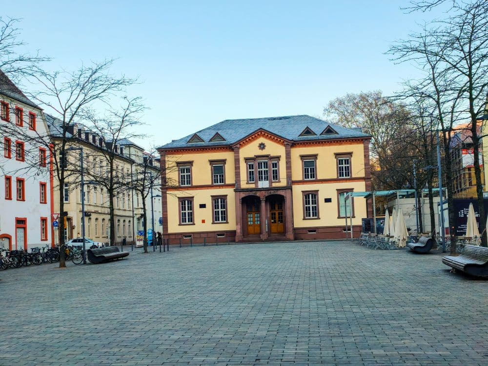 Ein kopfsteingepflasterter Platz mit blattlosen Bäumen umgibt ein historisches gelb-rotes Gebäude in Heidelberg mit einem zentralen Eingang und großen Fenstern; Sitzgelegenheiten im Freien und umliegende Gebäude vervollständigen die charmante Szene.