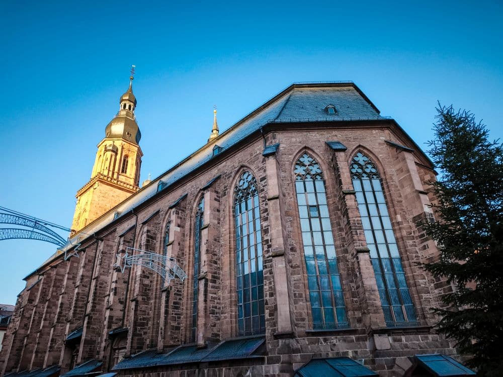 Eine große Steinkirche in Heidelberg mit hohen Bogenfenstern und einem Glockenturm steht vor einem klaren, blauen Himmel. Das Sonnenlicht beleuchtet einen Teil des Turms und einen Baum auf der rechten Seite.