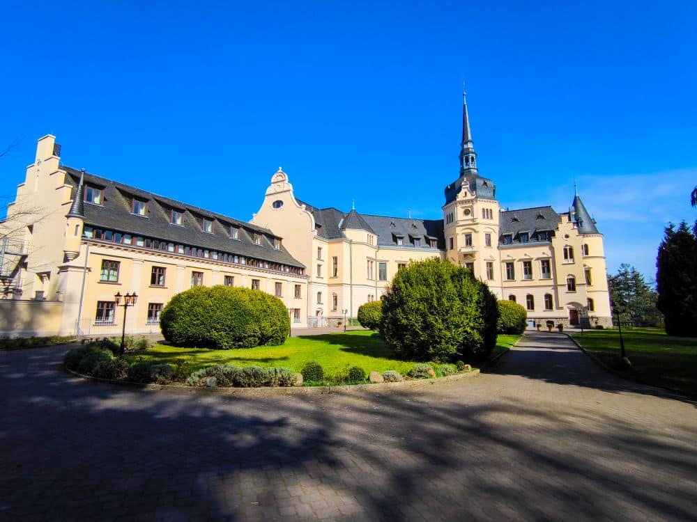 Ein prächtiges historisches Gebäude auf Rügen mit cremefarbenen Wänden, dunklen Schieferdächern und einem hohen zentralen Turm steht inmitten von gepflegten grünen Rasenflächen, Sträuchern und einem gepflasterten Gehweg unter einem klaren blauen Himmel.