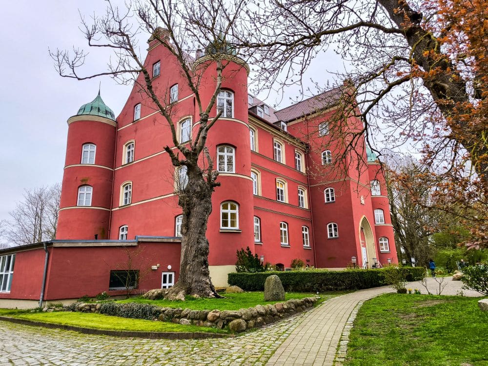 Ein großes rotes historisches Gebäude mit abgerundeten Türmen und vielen weiß verzierten Fenstern steht an einem bewölkten Tag an einem Steinweg auf Rügen, umgeben von Bäumen und Gras.