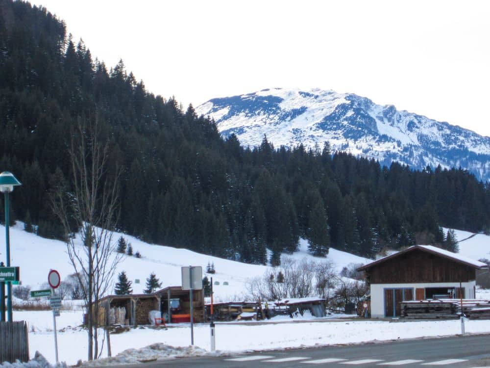 Eine verschneite Berglandschaft im Tannheimer Tal mit dichten Tannenwäldern, verstreuten Hütten und einer teilweise schneebedeckten Straße im Vordergrund bei bedecktem Himmel.