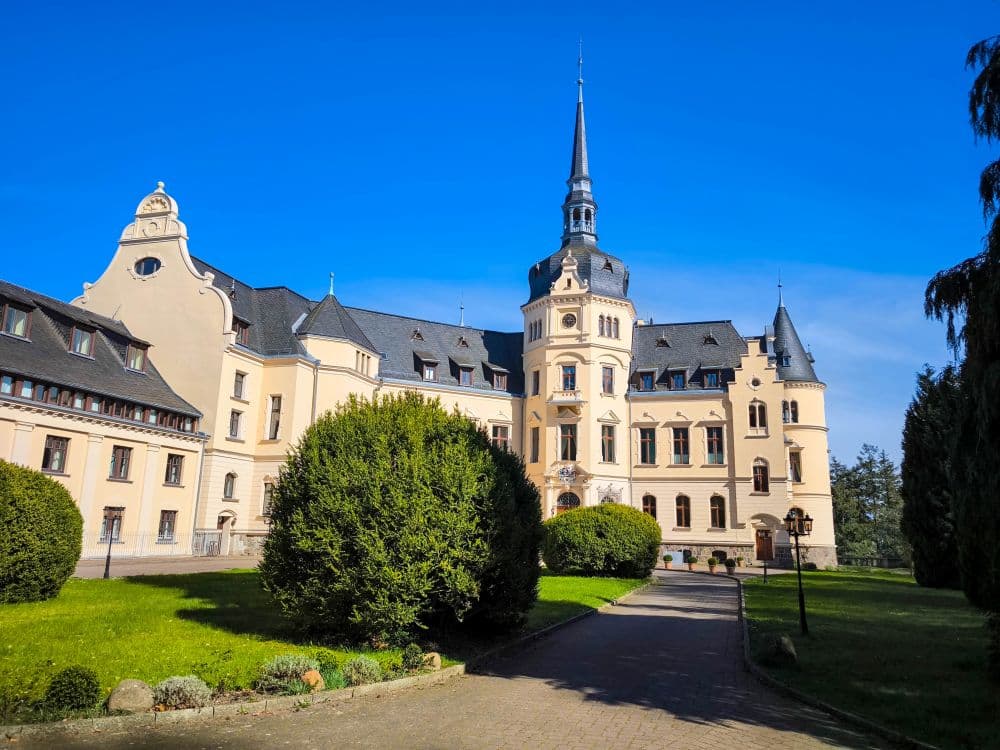 Unter blauem Himmel, umgeben von grünen Büschen und einem gepflegten Rasen, steht auf Rügen ein prächtiges, helles historisches Gebäude mit hohem Mittelturm und verziertem Dach.