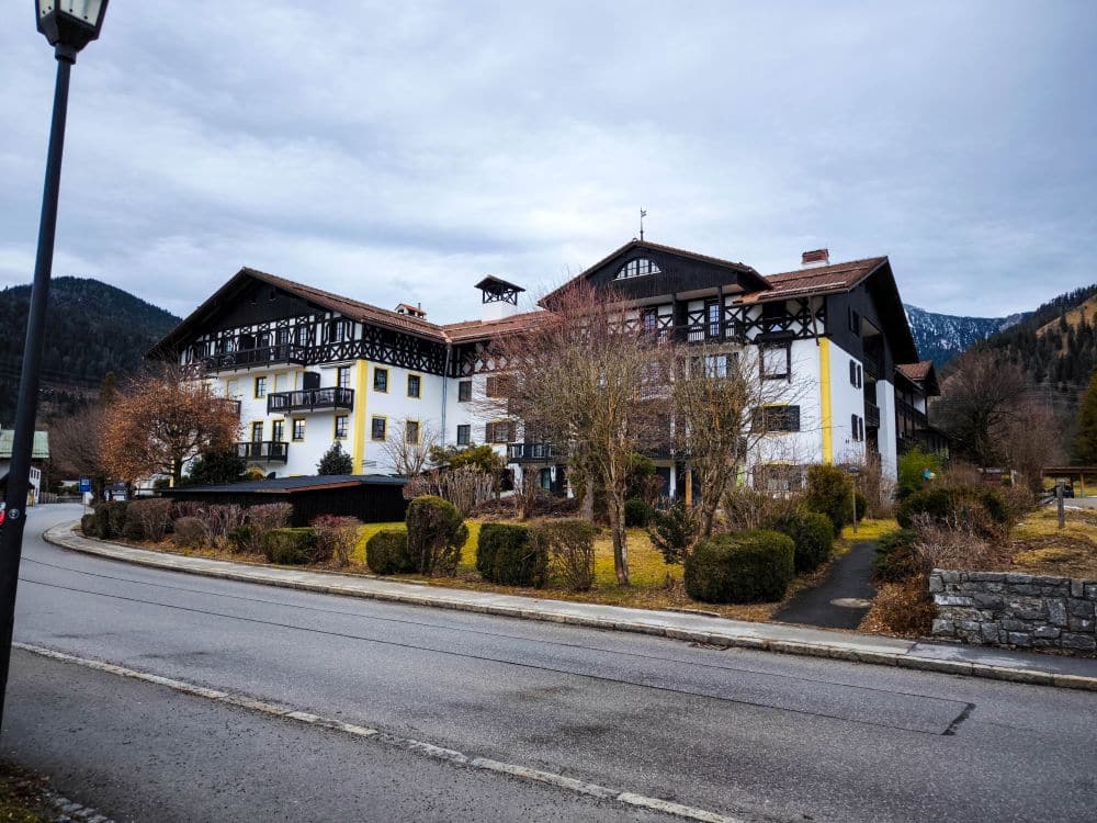 Ein großes, traditionelles Alpenhaus mit Holzbalkonen und weißen Wänden steht an einer ruhigen Straße in der Nähe des Walchensees, umgeben von gestutzten Sträuchern und blattlosen Bäumen, mit bewaldeten Bergen im Hintergrund unter einem bewölkten Himmel.