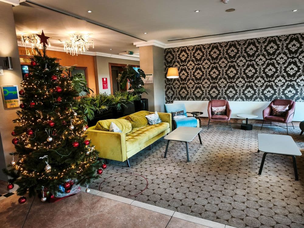 Eine gemütliche Heidelberger Hotellobby mit einem geschmückten Weihnachtsbaum, einem gelben Sofa, zwei Sesseln, gemusterten Tapeten und Couchtischen. Warme Beleuchtung und Pflanzen schaffen eine festliche und einladende Atmosphäre.