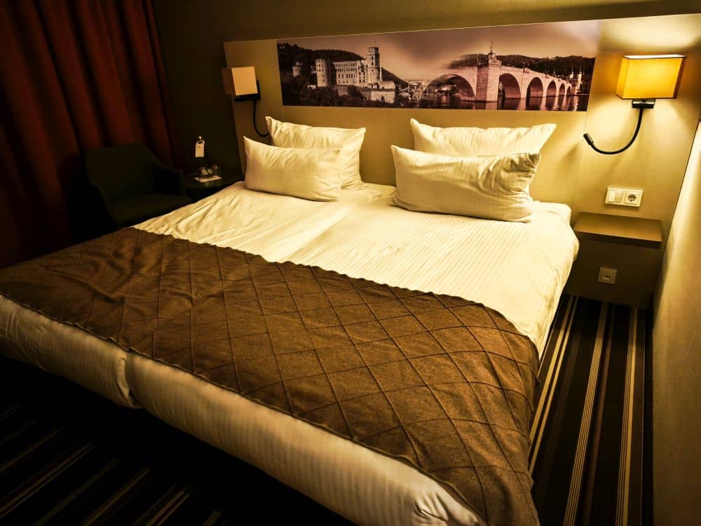 Ein ordentlich gemachtes Hotelbett mit weißer Bettwäsche, mehreren Kissen und einer braunen Decke; Nachttische mit Lampen auf jeder Seite und ein großes Schwarz-Weiß-Foto der Stadt Heidelberg über dem Kopfende.