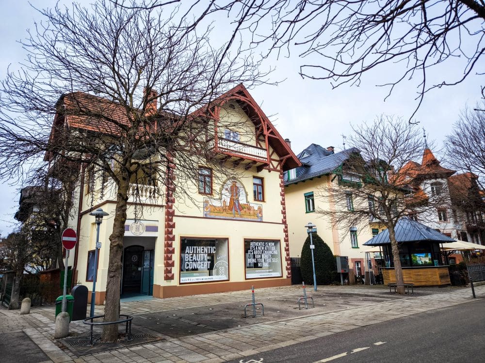 Eine Straßenecke mit einem großen, dekorativen Gebäude mit einem Wandgemälde und der Aufschrift Authentic Beauty Concept. Laublose Bäume säumen den Bürgersteig in der Nähe von leeren Fahrradständern und einem kleinen Sitzbereich im Freien, der an Szenen vom Starnberger See erinnert.