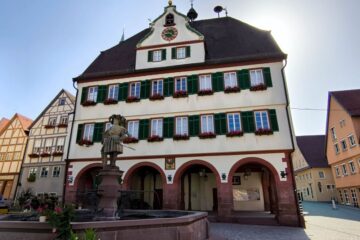 Ein historisches dreistöckiges Gebäude in Weil der Stadt mit grünen Fensterläden, Blumenkästen und einer Uhr an der Spitze; ein gewölbtes Erdgeschoss und eine Brunnenstatue stehen davor, Sonnenlicht scheint hinter dem Dach hervor.